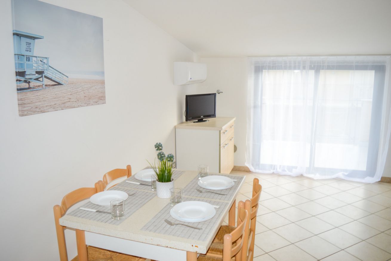 A dining area with a table set for four, a television on a stand, and a beach scene painting on the wall.