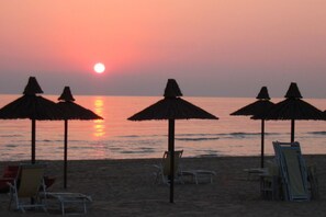 Ein Strand mit Sonnenschirmen und Liegen, ein Sonnenuntergang und ruhiges Wasser.