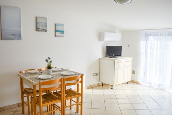 Un coin repas avec une table dressée pour quatre personnes, un meuble blanc sur lequel est posée une télévision à écran plat, et une fenêtre ornée de rideaux légers.