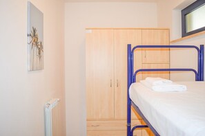 Un lit superposé avec un matelas blanc et une structure métallique bleue. On aperçoit une armoire en bois à deux portes et un petit tiroir. Un radiateur blanc est visible sur le mur de gauche, et un tableau encadré est accroché au-dessus.
