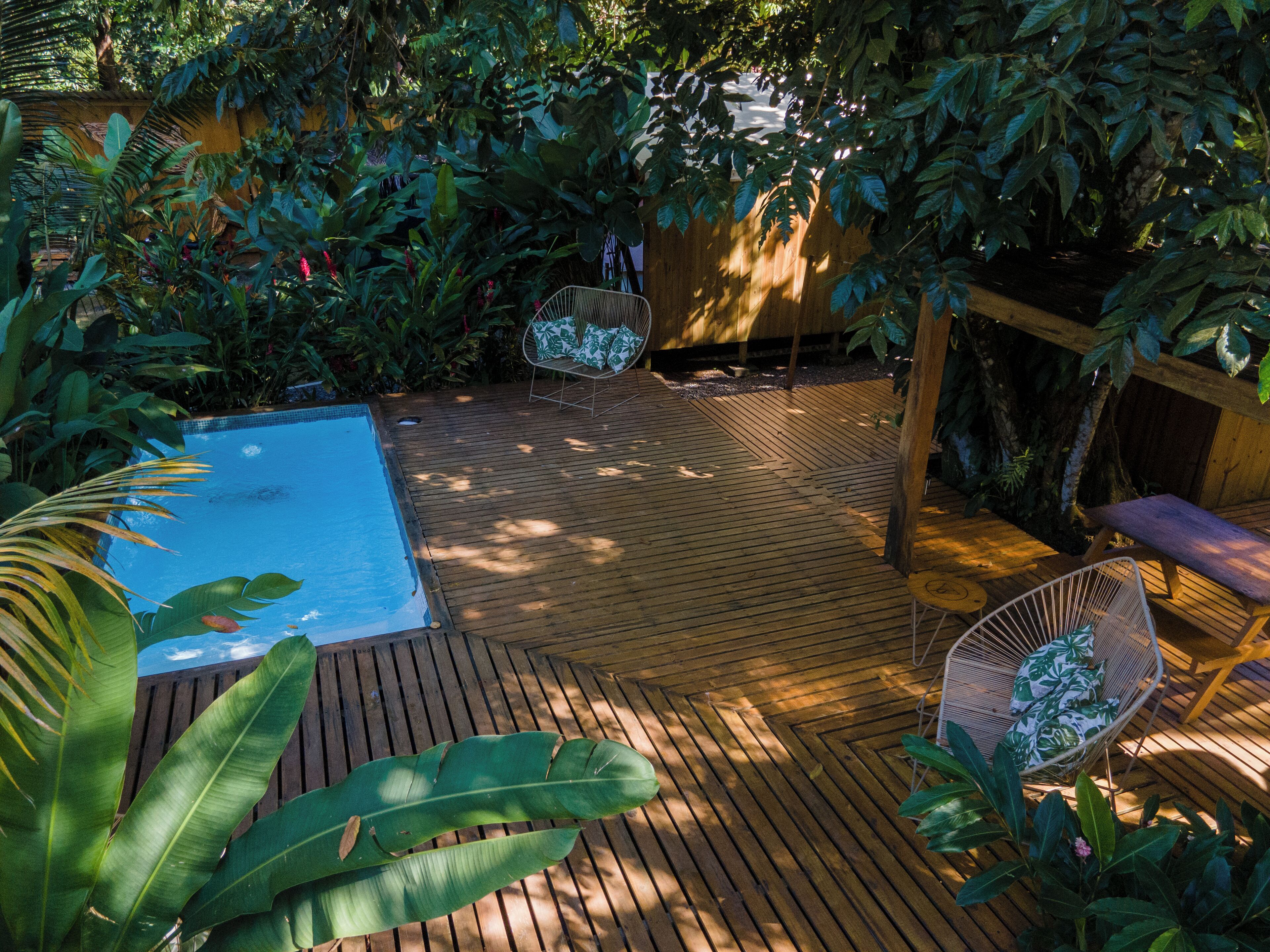 Une terrasse en bois avec une piscine, entourée d’une végétation luxuriante et d’une clôture en bois.