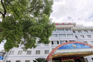A hotel with a large tree in the foreground, a blue awning with a logo, and a sign with Chinese characters. - Lg Hotel (Loudi)
