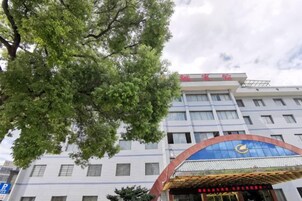 Et hotel med et stort træ i forgrunden, en blå markise med et logo og et skilt med kinesiske tegn.
