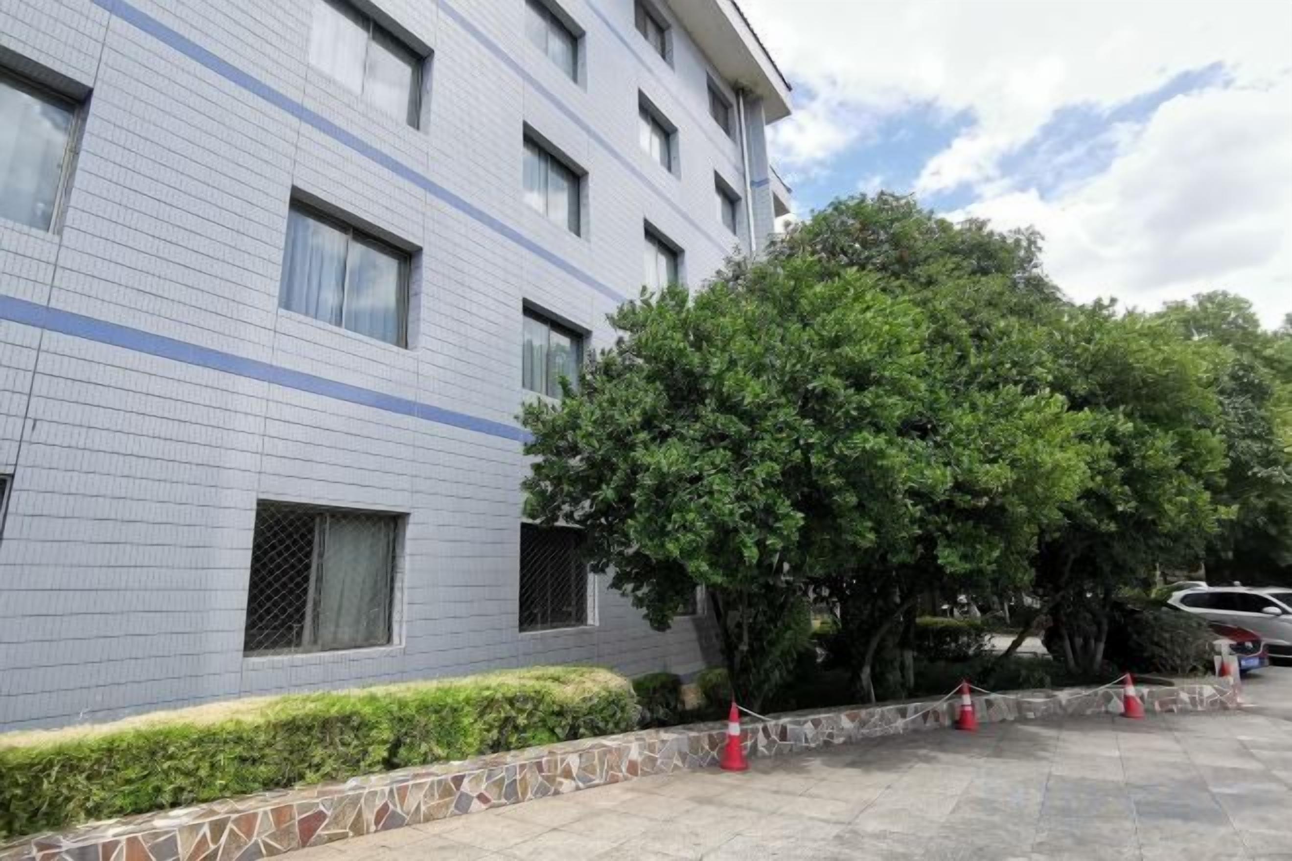 A multi-story building with a tiled facade, a tree-lined courtyard, and a parking area with a few cars.