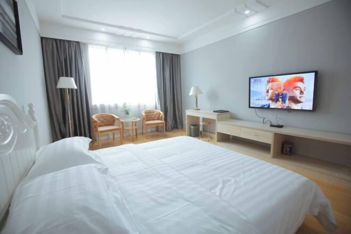 Ett hotellrum med en stor säng, ett skrivbord, två stolar, en tv på väggen och ett fönster med gardiner.