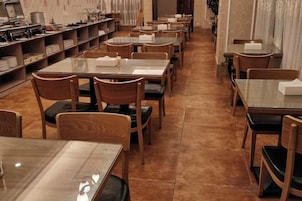 Un restaurante con varias mesas y sillas, suelo de baldosas y estanterías con objetos a la izquierda.