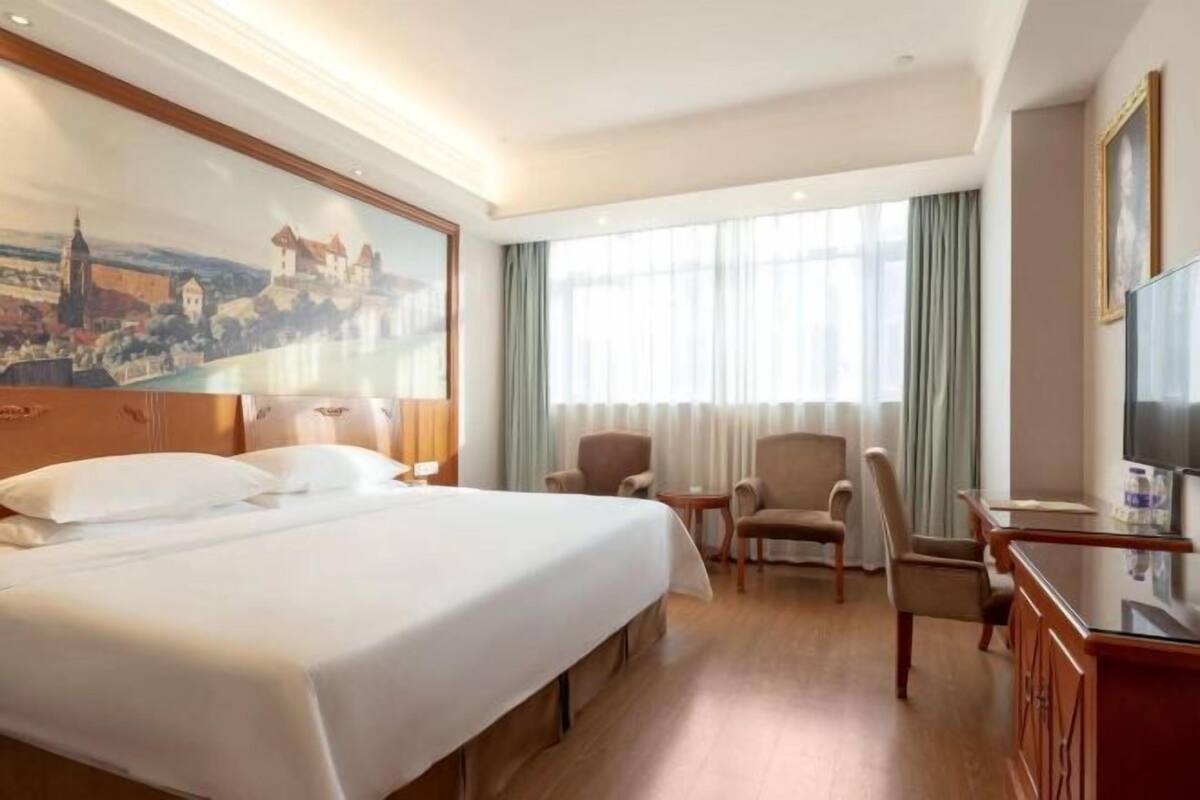 A hotel room with a large bed, two armchairs, a small table, a TV, and a painting of a castle on the wall.