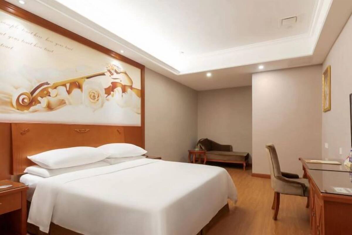 A hotel room with a large bed, a desk, a chair, and a painting on the wall.