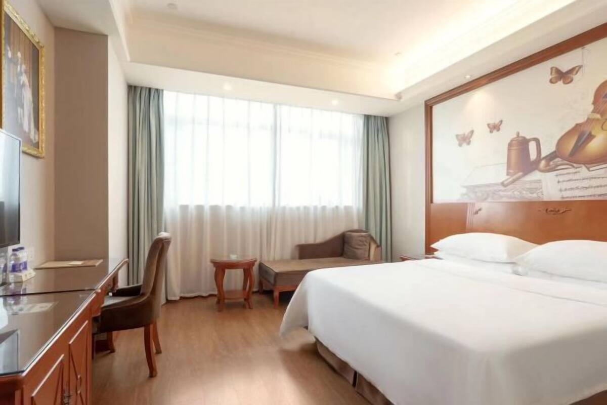 A hotel room with a bed, a desk, a chair, a small table, and a large window with curtains.