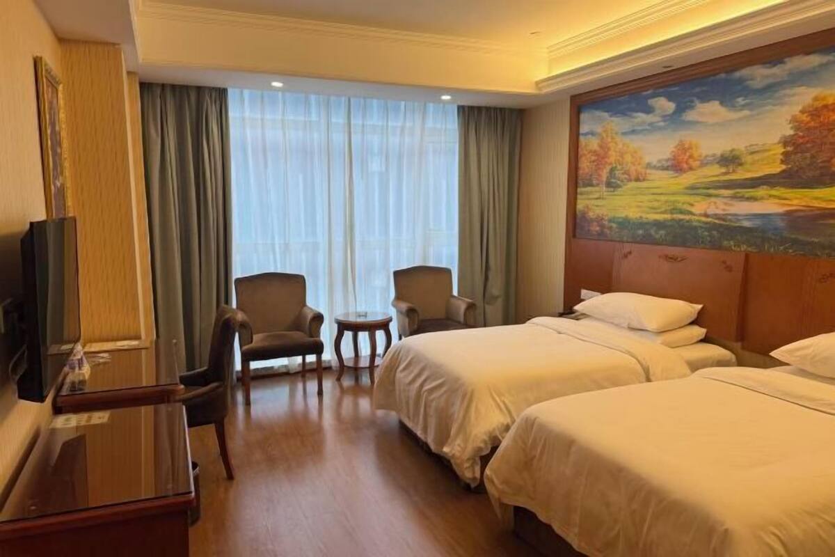 A hotel room with two beds, a desk, a chair, a small table, and a painting on the wall.