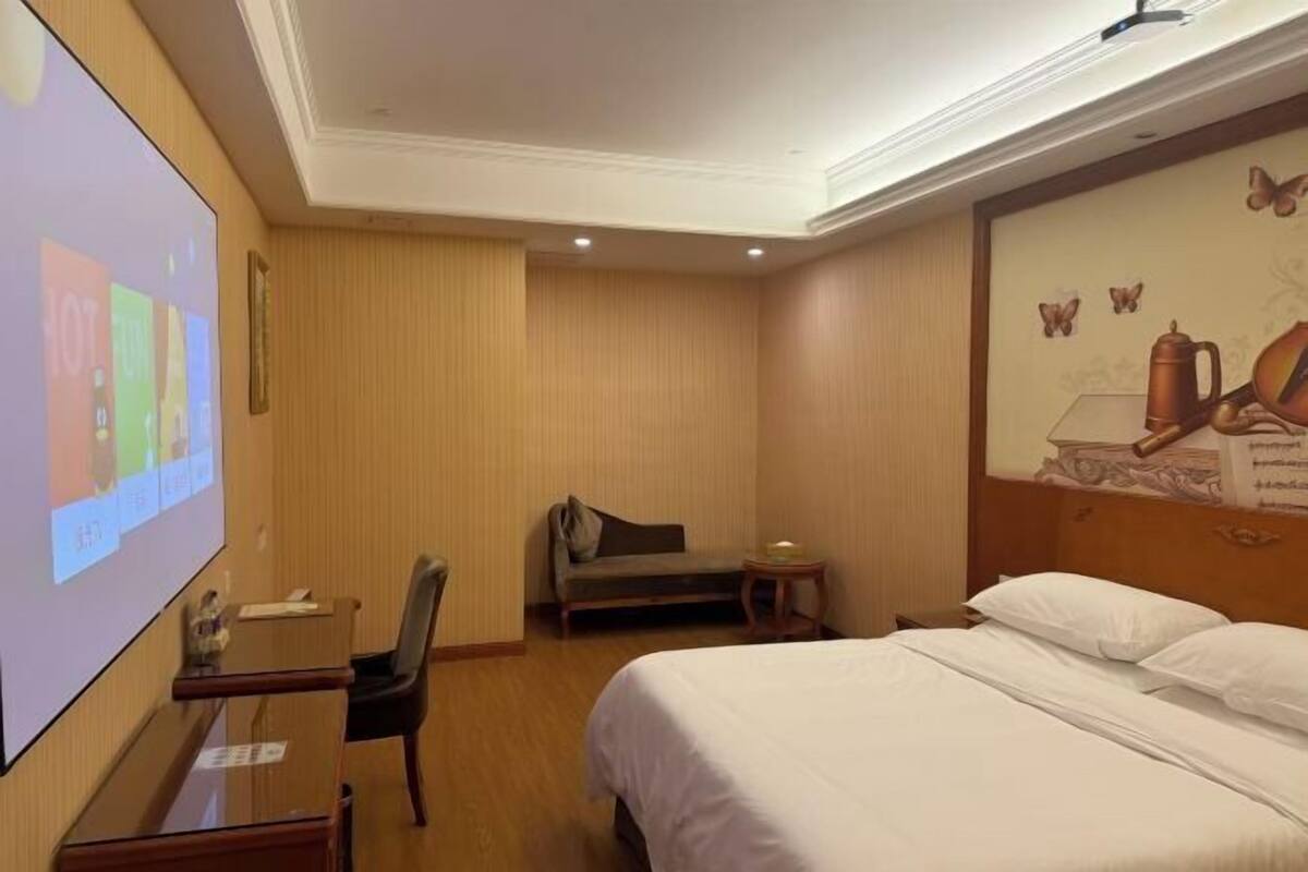 A hotel room with a bed, a desk with a chair, a small table, and a projector screen.