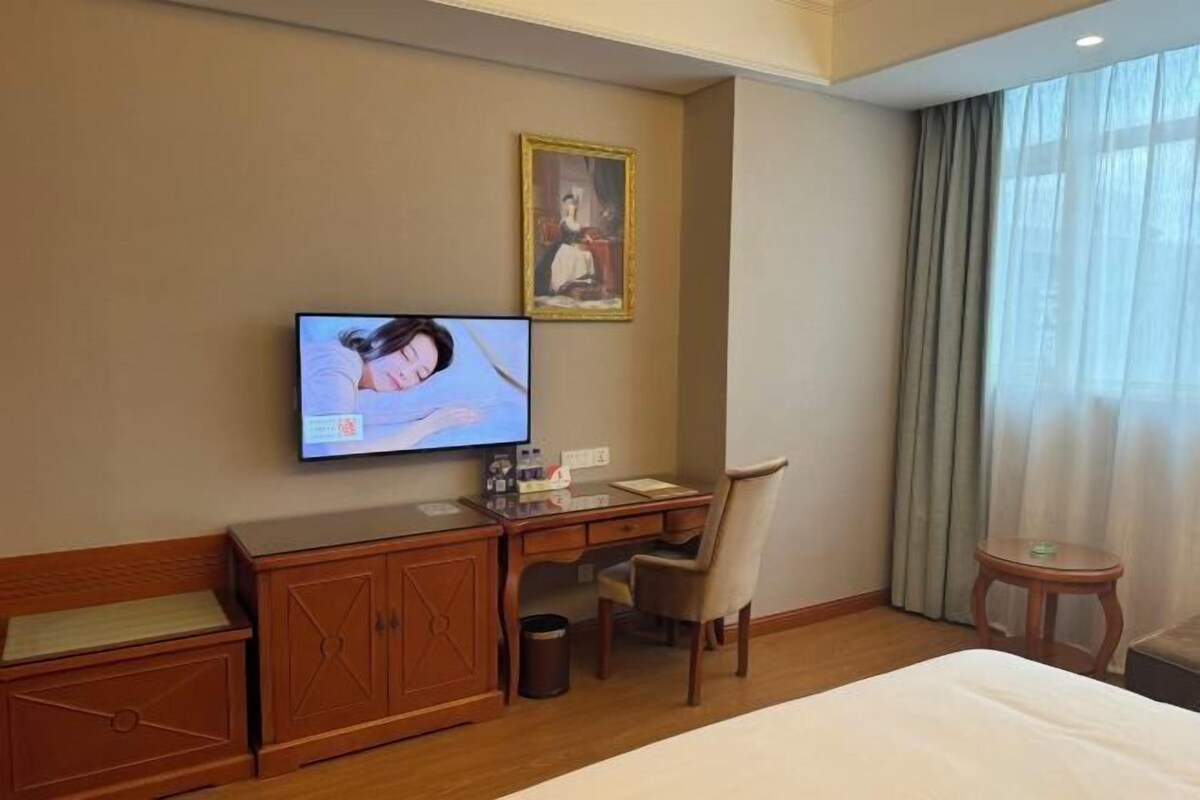 Habitación de hotel con cama, escritorio, silla, televisor, un cuadro y ventana con cortinas.