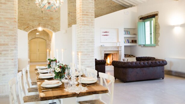 Un coin repas avec une longue table en bois dressée pour le repas, une cheminée et un canapé brun foncé dans une pièce aux murs en briques apparentes et ornée d’un lustre.