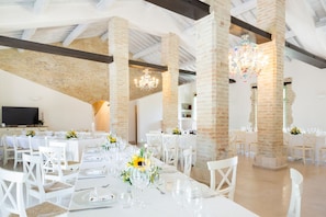 A spacious dining area with white tables and chairs, brick columns, and chandeliers.