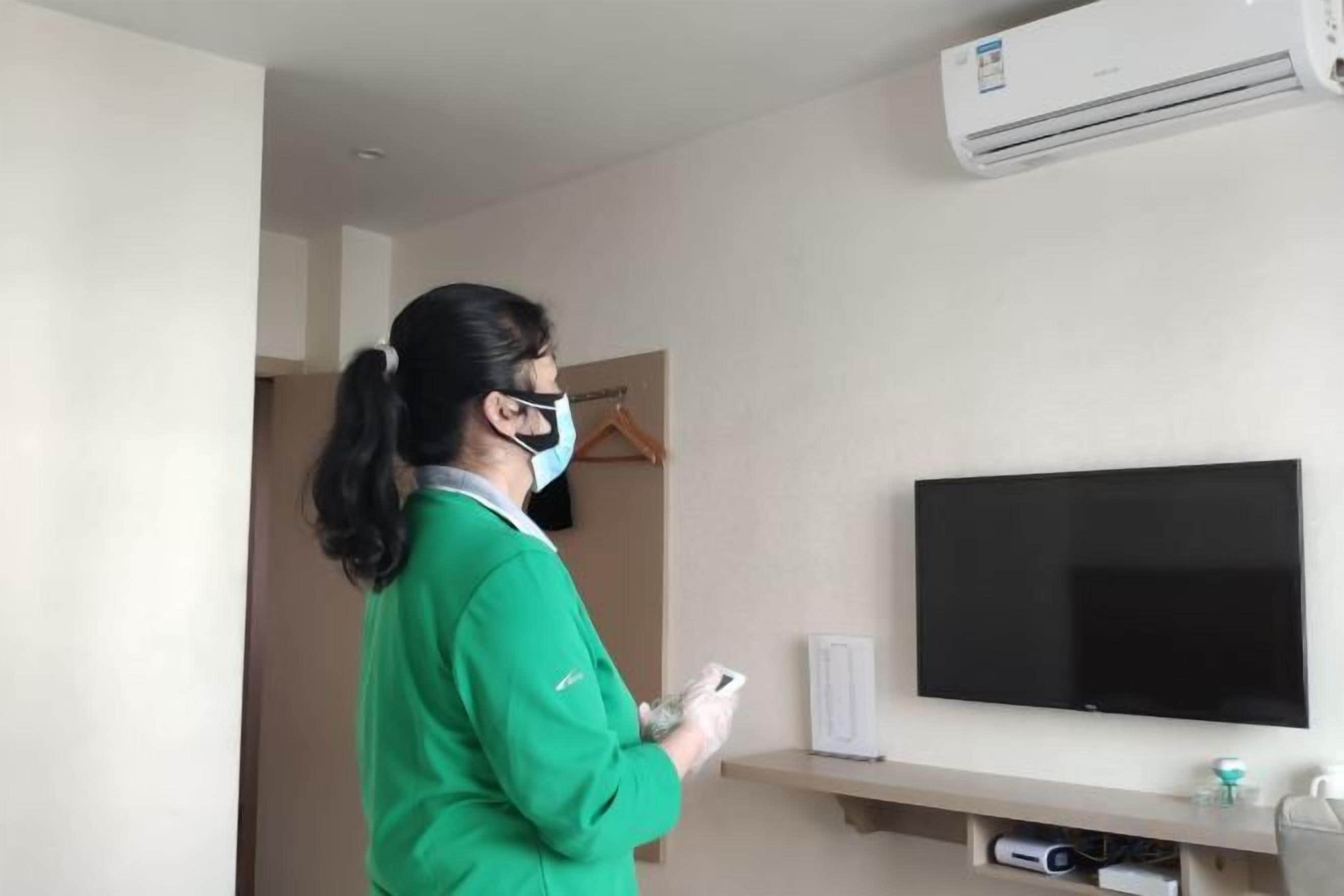A person in a green uniform holding a remote control in a room with a mounted flat-screen TV, a wall-mounted air conditioner, and a shelf with a small item and a container.