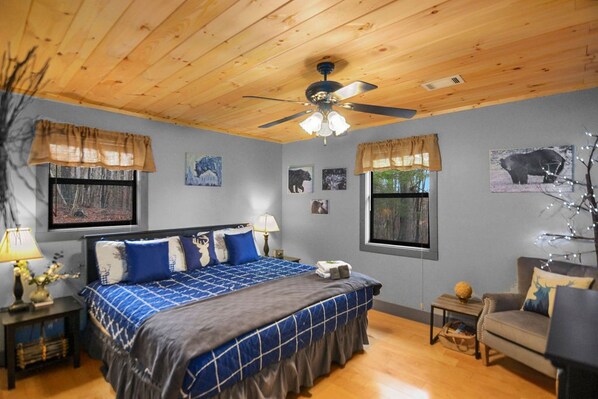 A bedroom with a wooden ceiling, a bed with blue bedding, a ceiling fan, and wall art including animal pictures.