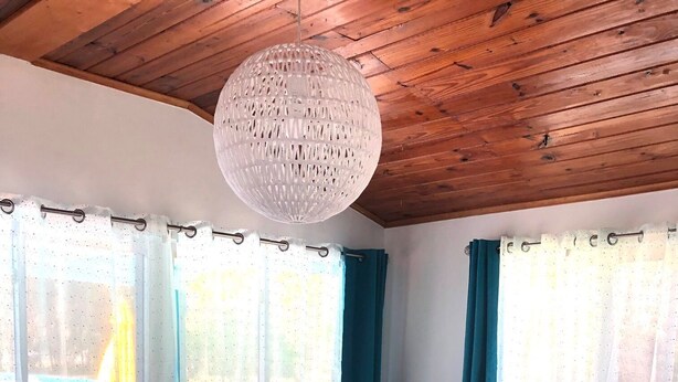 Un salón acogedor con techo de madera, una lámpara de araña redonda, un sofá con cojines de colores, un sillón mullido y una maleta antigua como mesa de centro.