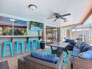 Un patio couvert aménagé avec des meubles en osier, des tabourets de bar bleus, un ventilateur de plafond et un écran de télévision.