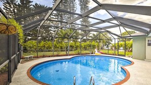 Una piscina techada con borde de ladrillo, rodeada por una estructura de metal y paneles de vidrio, ubicada en un área ajardinada con árboles y cielo despejado.