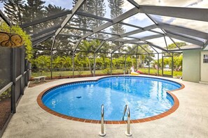 Ein überdachter Pool mit Ziegelkanten, umgeben von Metallrahmen und Glasscheiben, eingebettet in eine angelegte Grünfläche mit Bäumen und blauem Himmel.