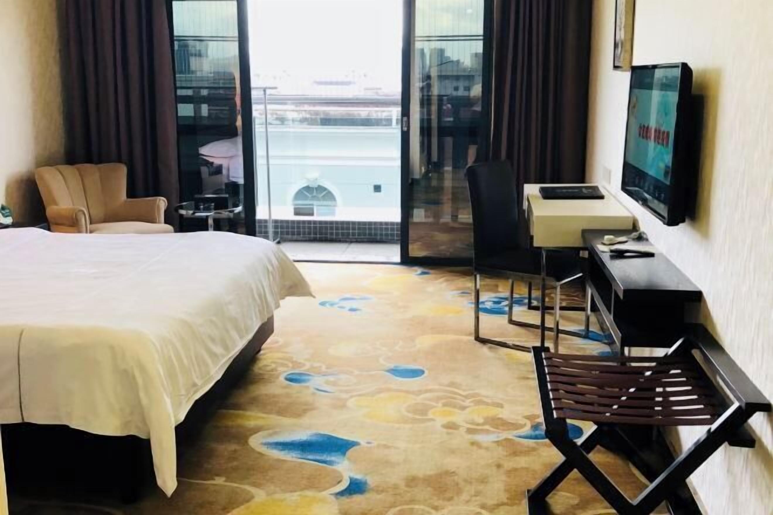 A hotel room with a large bed, a desk with a chair, a television, and a view through a glass door.