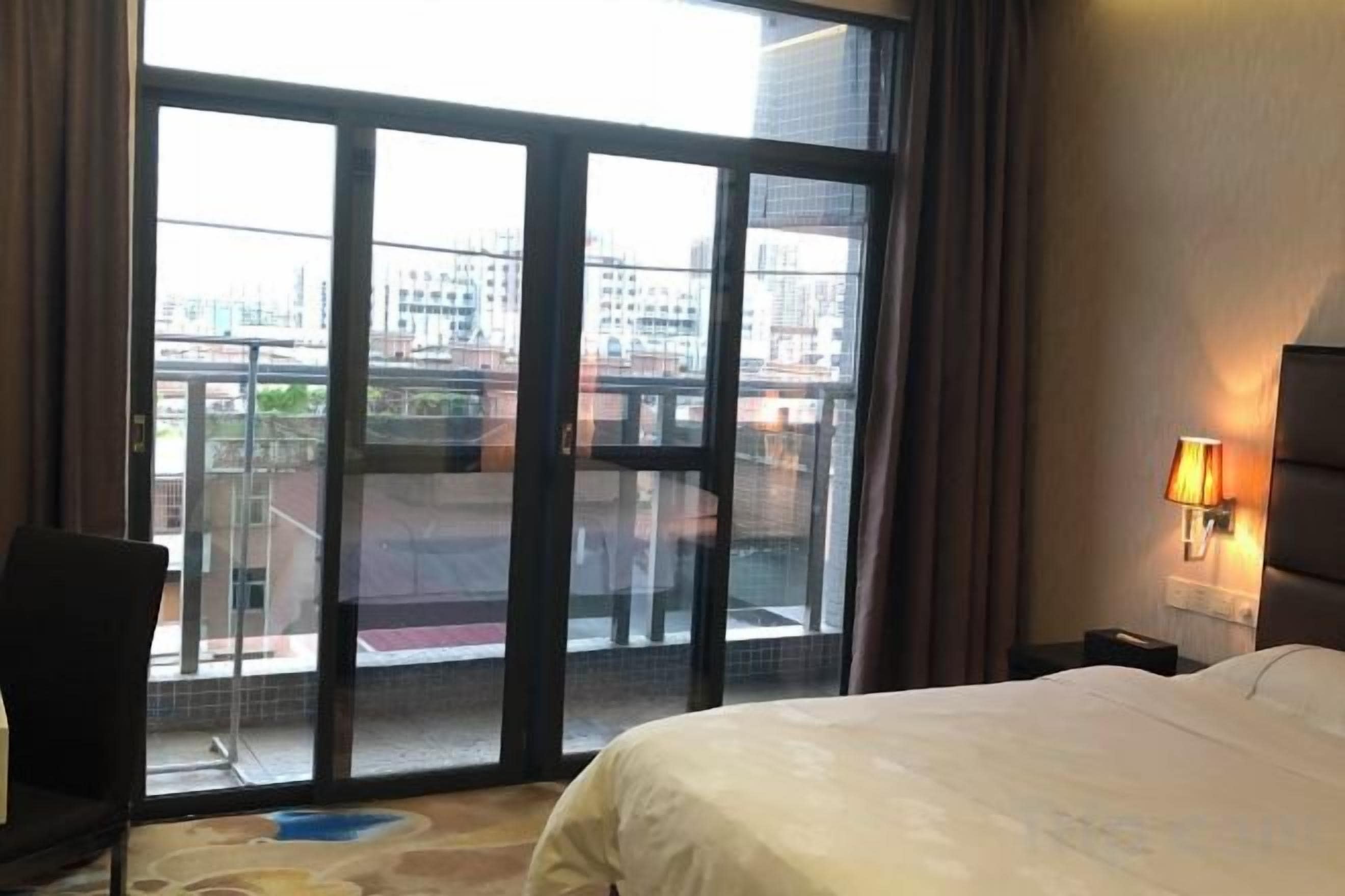 A hotel room with a large window, a bed, a chair, and a nightstand with a lamp.