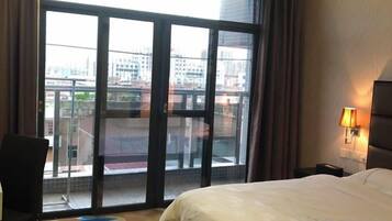 A hotel room with a large window, a bed, a chair, and a nightstand with a lamp.