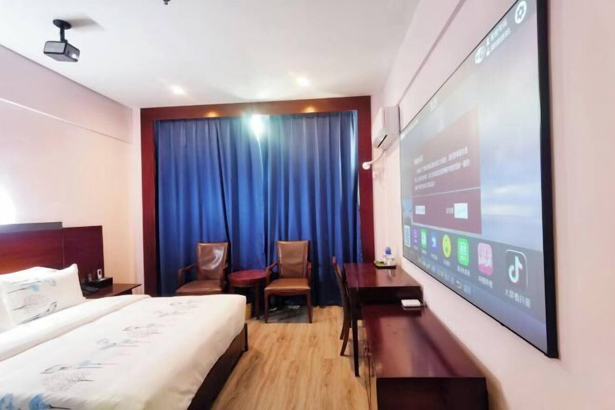 Hotelkamer met een bed, een bureau, twee stoelen, een projectiescherm en een groot raam met gordijnen.