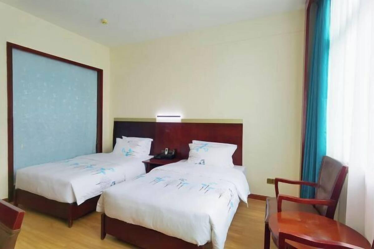 Een hotelkamer met twee enkele bedden, een houten hoofdeinde, een nachtkastje, een stoel en een raam met gordijnen.