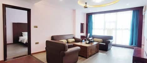 Habitación de hotel con cama, dos sofás, una mesa de centro y un ventanal con cortinas.
