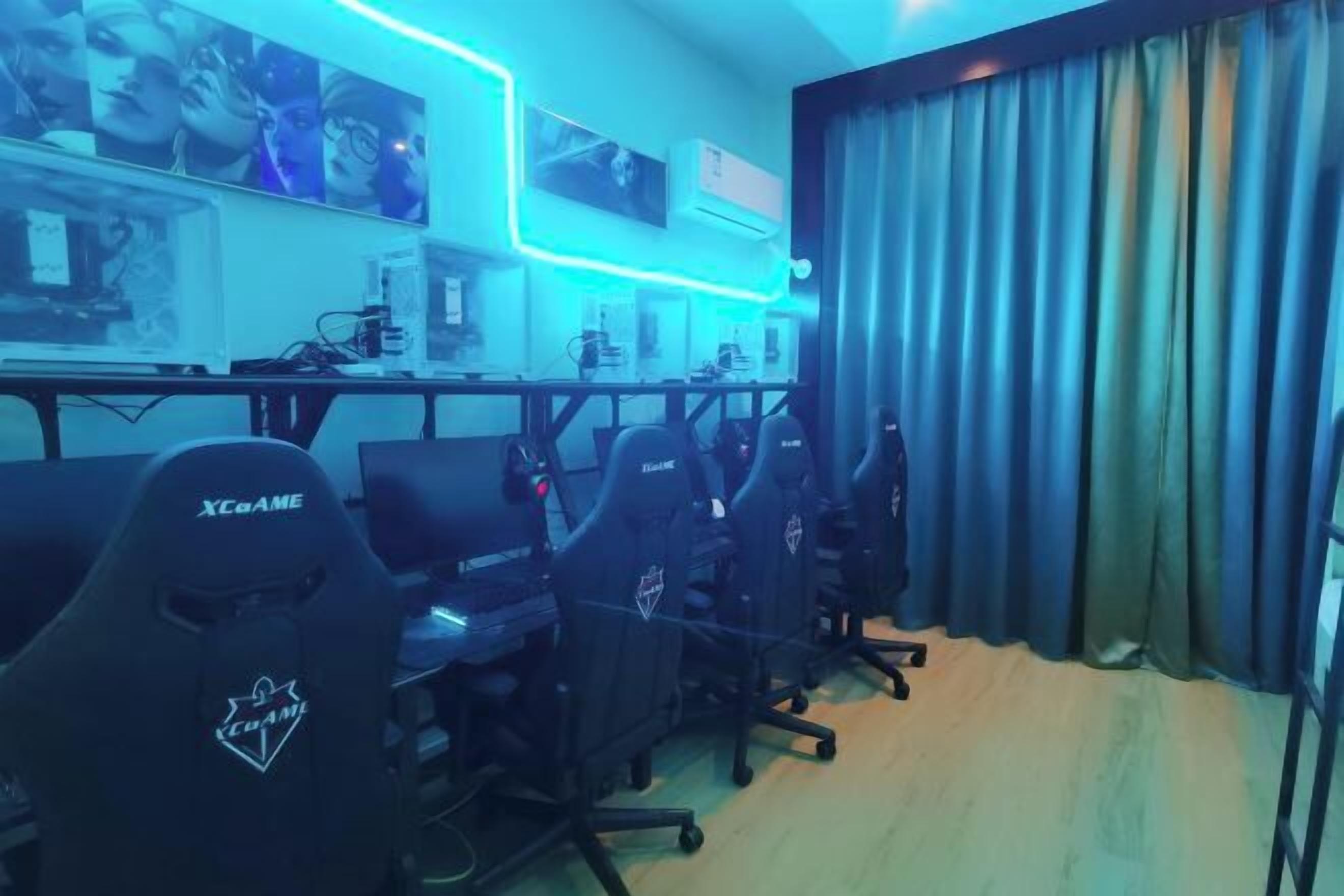 Una sala de juegos con varias sillas ergonómicas, un escritorio con computadoras y un monitor montado en la pared.