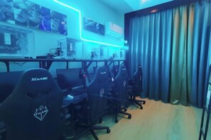 A gaming room with multiple gaming chairs, a desk with computers, and a wall-mounted monitor. - Deyuan Hotel (Shaoguan Nanhua Temple) (Shaoguan)