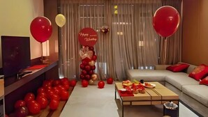 A modern living room with a sofa, a coffee table, and a TV. There are balloons and a 'Happy Wedding' sign.