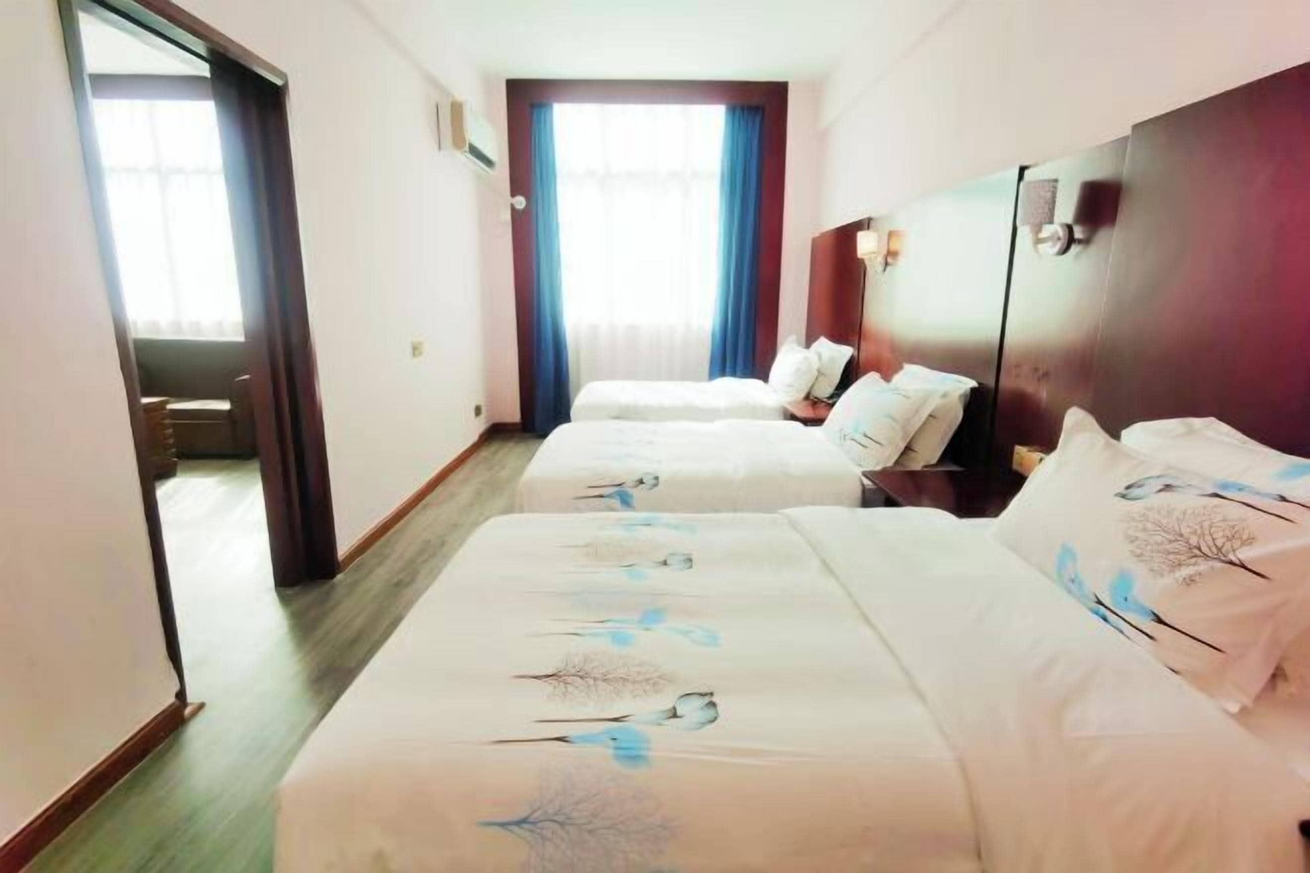 Une chambre d’hôtel avec deux lits, une tête de lit en bois, une fenêtre avec des rideaux et une porte donnant sur une autre pièce.