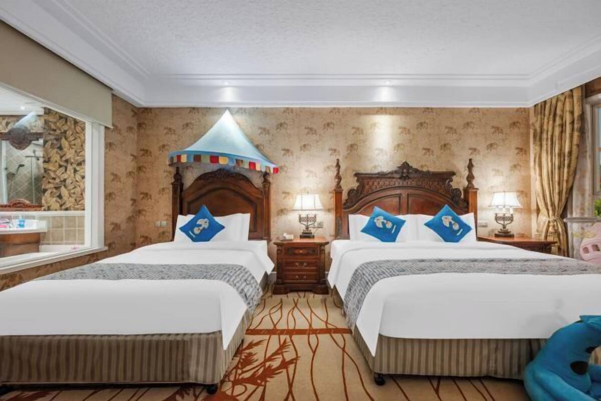 A hotel room with two beds, a wooden headboard, a patterned carpet, and a window with a view of a building.