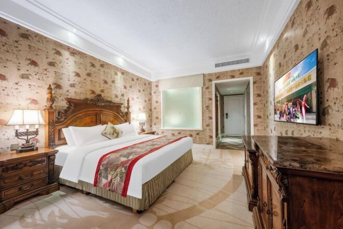 A hotel room with a large bed, wooden headboard, a nightstand with a lamp, a dresser, and a television on the wall.