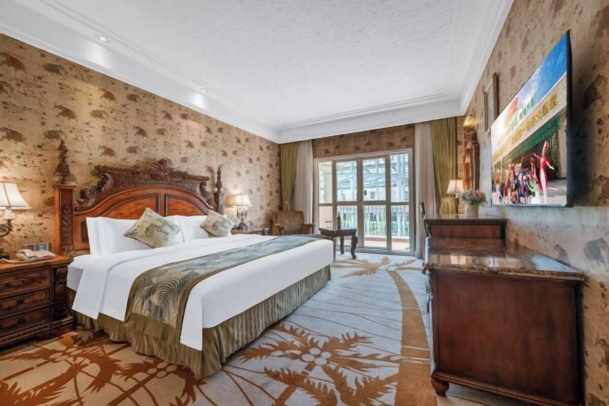 A hotel room with a large bed, wooden headboard, a nightstand, a desk, a chair, and a large window with a view.