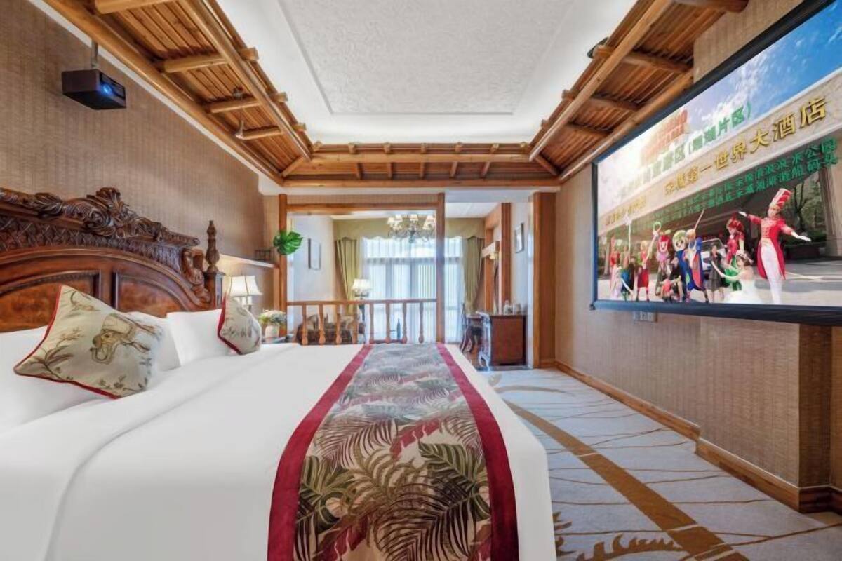 A hotel room with a large bed, a wooden headboard, a television screen displaying a performance, and a view of the outside.