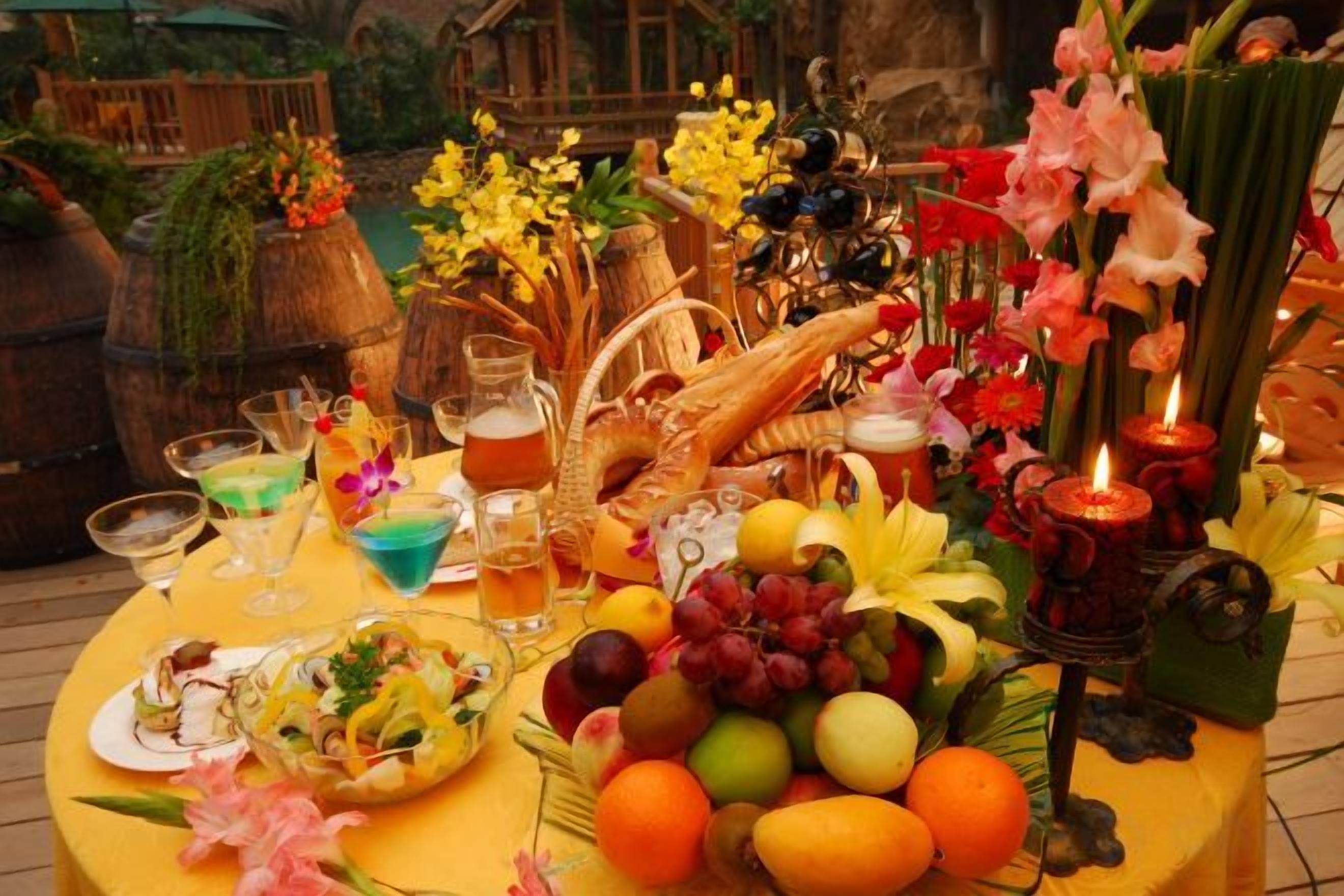 Una mesa con frutas, flores y bebidas, ubicada al aire libre con mobiliario de madera y barriles al fondo.