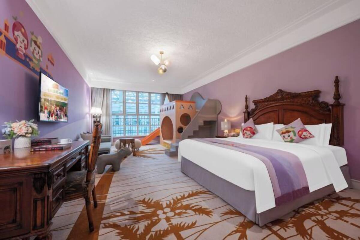 A hotel room with a large bed, a desk, a chair, a TV, and a play area with a slide.