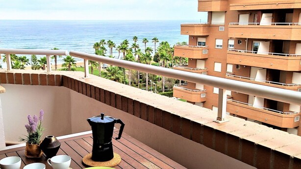 Een balkon met een houten tafel, een koffiezetapparaat, bekers en een bord. In het zicht: een gebouw en palmbomen.