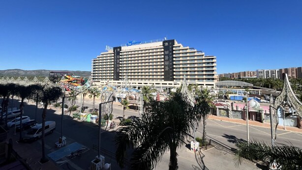 Un grand hôtel à plusieurs étages, doté de balcons et entouré de palmiers, avec un espace de stationnement à proximité.