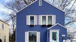 Ett tvåvåningshus med en blå fasad, vita fönsterluckor och en veranda framför huset med trappsteg och ett räcke.