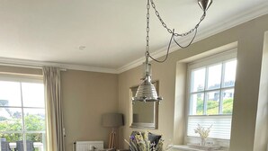 A dining area with a round table, four chairs, a hanging light fixture, and a window with blinds. - Heefkant HK/01 - Heefkant (Sylt)