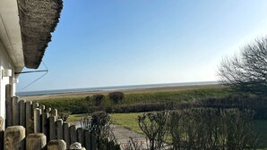 A coastal view from a balcony with a thatched roof, a wooden fence, and a grassy landscape leading to the sea. - Heefkant HK/01 - Heefkant (Sylt)