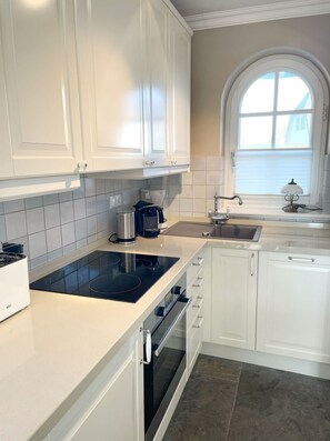 A modern kitchen with white cabinets, a black induction cooktop, a built-in oven, a window with a view, and a sink with a faucet. - Heefkant HK/01 - Heefkant (Sylt)