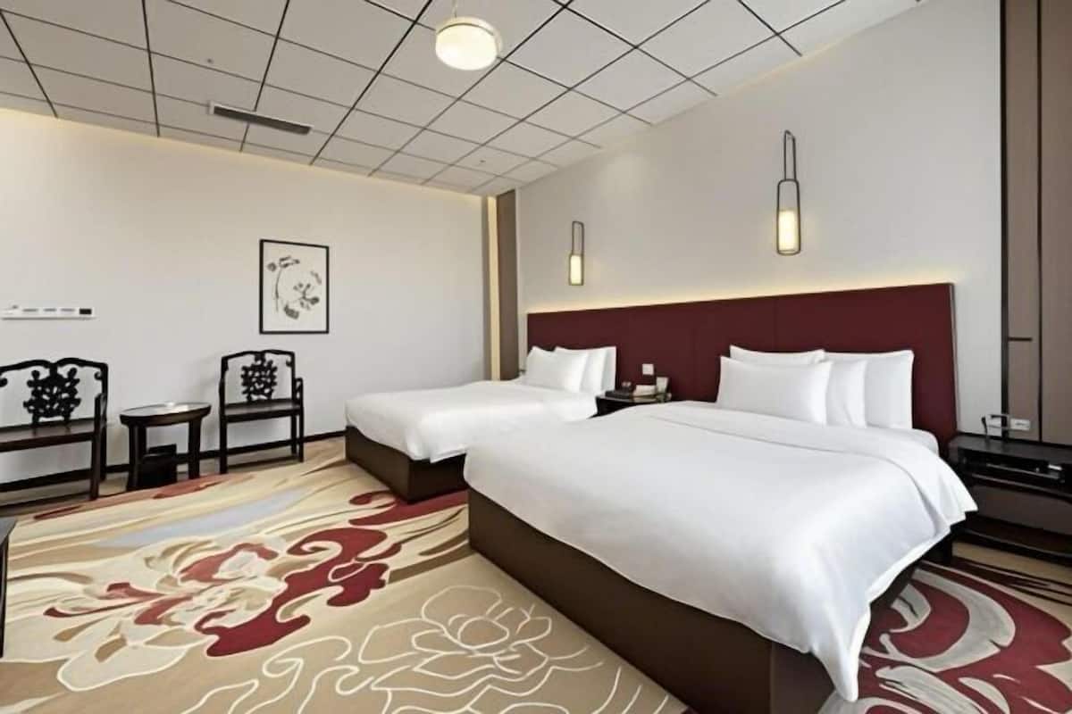 Hotellihuone, jossa on kaksi sänkyä, punainen pääty, kuvioitu matto ja seinään kiinnitetyt valaisimet.