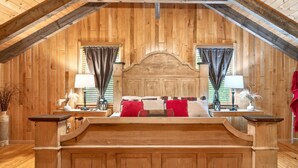 A cozy attic bedroom with wooden walls and a sloped ceiling. It features a central bed with red and white bedding, two bedside tables with lamps, and a large wooden headboard.