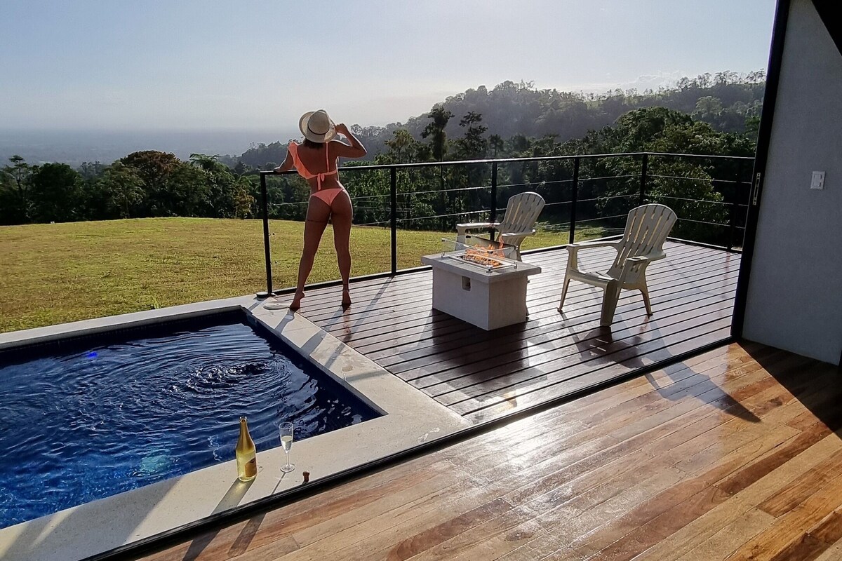 A modern outdoor pool area with a wooden deck, a fire pit, and a view of a lush landscape.
