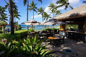 Een terras buiten met parasols, tafels en stoelen, met uitzicht op een strand met palmbomen en een constructie met rieten dak.
